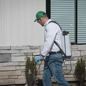 A First Line Pest Defense technician applying pest control treatment to the exterior perimeter of a residential house.