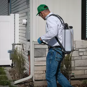 A First Line Pest Defense technician applying pest control treatment to the exterior perimeter of a residential house.
