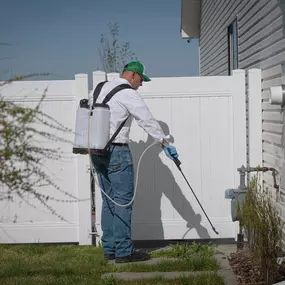 A First Line Pest Defense technician applying outdoor pest control treatment near a fence and building exterior.