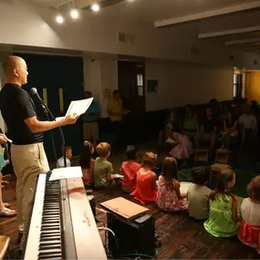A lively music class setting with instructors addressing a group of children seated on the floor, reflecting the engaging learning atmosphere of Musicolor Method NYC.
