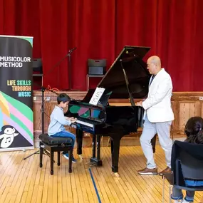 A young student is playing the piano on stage with an instructor beside them, creating a warm performance moment that reflects Musicolor Method NYC’s supportive approach to piano lessons NYC.