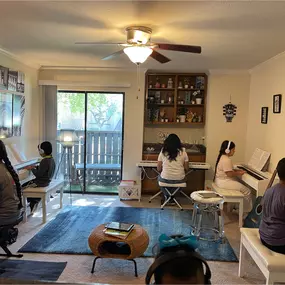 This photo shows a well-organized, cozy piano lesson studio where multiple students are engaged in their music practice. Each student is seated at their digital piano, wearing headphones, creating a focused and distraction-free environment for learning. The room is thoughtfully designed with soft lighting, modern decor, and wall artwork, including an artistic Eiffel Tower-themed gallery and a creative painting on the right side.

The studio’s open, inviting space includes a bright window and sli