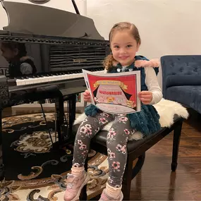 This photo captures a heartwarming and proud moment at a music studio. A young girl sits happily on a piano bench, holding a colorful 