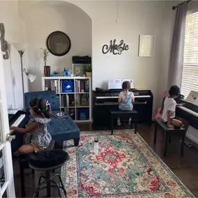 This photo showcases a bright and inviting piano studio where three young students are actively engaged in their piano lessons. The room is thoughtfully arranged with three digital pianos, each with its own space, allowing students to focus independently while learning. The children are seated comfortably, wearing headphones to create a distraction-free, immersive learning experience.

The studio’s design is both functional and warm. A stylish “Mu$ic” wall decoration adds a creative touch while 