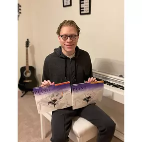 This photo features a smiling student proudly showcasing two Piano Adventures lesson books while seated at a sleek white digital piano. The student’s joyful expressions and relaxed posture reflect a sense of accomplishment and excitement about their musical journey.

In the background, a black acoustic guitar rests against the wall, along with musical-themed decorations that enhance the room’s creative and inspiring vibe. The clean, organized studio space creates a calm and welcoming environment