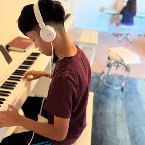 This photo captures a focused moment at Baney Jones Music Studio, where a young student is deeply engaged in piano practice. Wearing white Sony headphones, he is fully immersed in his music, concentrating on the keys of a sleek white digital piano. His posture and expression show dedication, discipline, and a love for learning.

In the background, another student also practices at a separate piano, reinforcing the studio’s engaging and structured learning environment. The warm lighting, soft blu