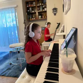 This photo captures a focused and immersive Baney Jones Music Studio music lesson. Young students are deeply engaged in their piano practice, Wearing Sony headphones. They concentrate on their pieces and refine their skills in a structured, distraction-free environment.

In the background, another student is multitasking, alternating between piano and guitar, showcasing the studio’s versatile approach to music education. The setting is warm and inviting, with an organized learning space and a co