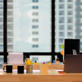 Modern office desk setup with laptops, mugs, and stationery, overlooking a city skyline through large windows.