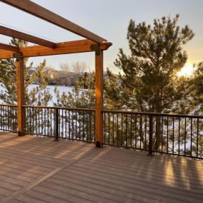 A sunset picture of a composite deck. The deck has Deckorators ALX Contemporay Brown railing with a drink rail (deck board) mounted on top. A redwood pergola is also off to the side with black decorative hardware.