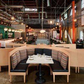 This image highlights the modern, industrial design of Punch Bowl Social, featuring a spacious booth with colorful cushions and an open bar area. The large windows and warm lighting create a welcoming atmosphere perfect for social gatherings.