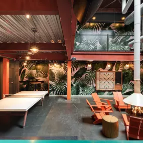 This image shows the vibrant lounge area at Punch Bowl Social, featuring table tennis, tropical-themed wallpaper, and a cozy fireplace surrounded by orange Adirondack chairs. The space provides a fun and relaxed atmosphere for guests.