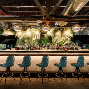 This photo highlights the bar at Punch Bowl Social, with blue cushioned bar stools and a tropical-themed wall. The modern lighting and decor create a stylish yet inviting ambiance.