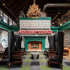 This image showcases the Holiday Lodge area at Punch Bowl Social, featuring high-backed chairs, a cozy fireplace, and a 'Holiday Lodge' neon sign. The space's warm ambiance makes it perfect for a relaxing social experience.