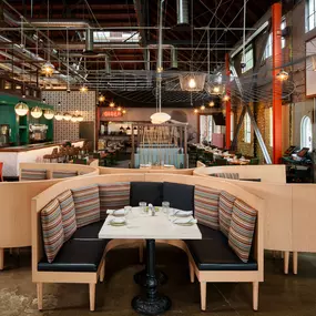 This image highlights the modern, industrial design of Punch Bowl Social, featuring a spacious booth with colorful cushions and an open bar area. The large windows and warm lighting create a welcoming atmosphere perfect for social gatherings.