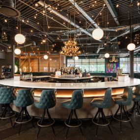 This photo showcases a circular bar at Punch Bowl Social Atlanta, with teal bar stools and modern hanging lights. The bar area is inviting and vibrant, perfect for social gatherings.