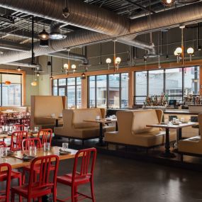 This image showcases the modern and vibrant dining area at Punch Bowl Social Atlanta, featuring red chairs, tan booths, and large windows that let in plenty of natural light. The space is both spacious and cozy, perfect for social dining.