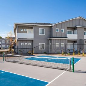 View of gated tennis/pickleball courts with grey shiplap style buildings in the background