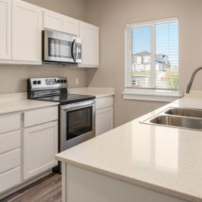 Neutral colored kitchen with refrigerator, stove with microwave above, white cabinetry, across from countertop with dual sided sink and gooseneck faucet