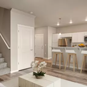 View from living room into kitchen with contemporary coffee table, plank flooring, view of barstool counter seating across from the kitchen area, and side view of stairs leading up to bedrooms