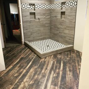 A corner shower adorned with barnwood-style gray tiles. The white and black design accents add a visual contrast against the tiles. The surrounding barnwood floors create a cozy atmosphere, blending modern and rustic elements seamlessly.