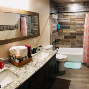 A guest bathroom featuring a double vanity with a granite countertop. The wood-toned tiled shower adds warmth to the space. The double vanity, wood-toned tiles, and granite countertop creates a modern and inviting atmosphere in this guest bathroom.