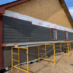 New home under construction in the siding phase, with scaffolding in place. Gray wood siding with red trim adds a stylish touch to the exterior, showcasing progress in the construction process.