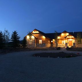 Newly completed modern Western home with rock and wood siding, illuminated by exterior lights at night.