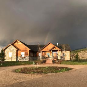 Modern home with rock and wood siding. Circular driveway. Overcast sky with a vibrant rainbow behind the house, adding an enchanting touch to the scene.