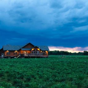 Newly completed home with cedar siding, situated in a prairie landscape. The evening scene features an overcast sky at sunset, with exterior lights illuminating the house, creating a cozy and inviting atmosphere.