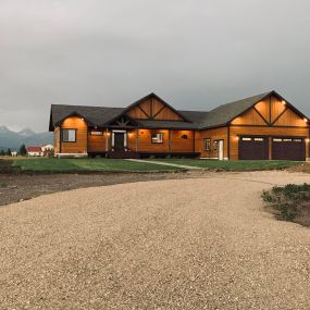 Evening view of a newly constructed home with cedar siding. Exterior lights illuminate the house, creating a warm ambiance. The magnificent Grand Tetons serve as a breathtaking backdrop, adding to the natural beauty of the scene.