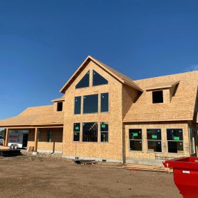 Under-construction new home with large windows, high peaked ceiling, and no siding or shingles. Blue skies and snow-covered ground complete the scene.