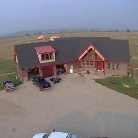 Arial photo of a newly constructed modern Western home on a spacious prairie plot. The house features grey siding, red trim, and a 3-car attached garage, blending harmoniously with the vast natural surroundings.