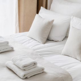 Hotel & Hospitality Laundry Services in Largo, FL. Cropped view of white bedclothes and towels on bed in comfortable hotel room, hypoallergenic pillows, personal comfort idea, bedding concept, bathroom details.