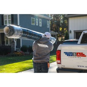 A worker from USA Flooring carrying a rolled-up carpet on his shoulder, walking towards a bluish house in the background.