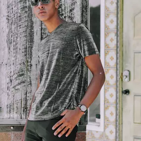 This image, developed by Sapien Designs Marketing Digital SEO, showcases a person standing against a rustic wooden wall, wearing a grey t-shirt and black pants, with a stylish watch on the wrist.