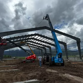 The image shows the construction of a steel structure for a horse arena by Steel Concepts, Inc. The structure is being erected with the help of heavy machinery, including a blue crane and a red lift. The steel framework is partially completed, with beams and columns in place. The construction site is located in Swan Valley, ID, and the project covers 17,600 square feet. The image captures the progress of the building erection under a cloudy sky, highlighting the scale and complexity of the proje