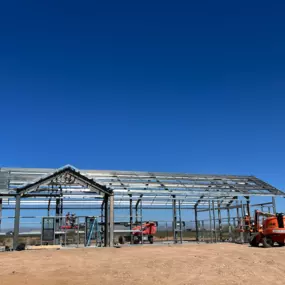 The image shows a steel structure under construction, which is part of a project by Steel Concepts, Inc. The structure is a Barndominium with a total area of 4,500 square feet, located in Wittmann, AZ. The construction site features a clear blue sky and a desert landscape in the background. The steel frame of the building is partially erected, with workers and construction equipment visible on-site. This project is categorized as residential and is specifically for the Huskinson Riding Arena.