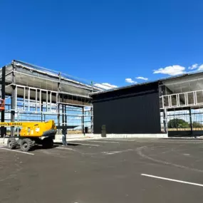 A partially constructed prefabricated steel frame commercial building in Thornton, ID. The structure features exposed steel framing, black metal siding, and wood paneling. The surrounding area includes a parking lot, dirt ground, and a clear blue sky.