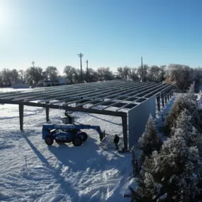 The image shows a steel structure under construction in a snowy landscape. The structure is a large, rectangular framework with steel beams and columns, partially covered with metal siding on one side. A blue construction vehicle with 