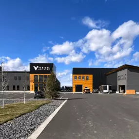 A newly constructed commercial office building with a prefabricated steel frame structure, featuring a modern design with black, gray, and wooden elements. The surrounding area includes a parking lot, landscaping with rocks and grass, and a clear blue sky with scattered clouds.