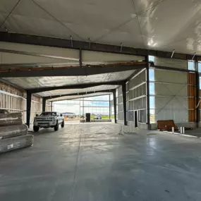 Interior view of a prefabricated steel frame commercial building under construction. The space features steel beams, insulation, and unfinished walls. A truck is parked inside, with construction materials stacked on one side and an open view to the outside at the far end.