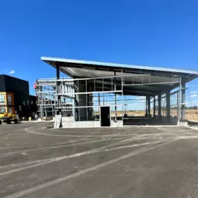 An exterior view of a 7,600 sq. ft. prefabricated steel frame commercial building under construction in Thornton, ID. The structure showcases exposed steel beams and an angled roof, with surrounding construction materials and equipment visible. Nearby is a completed building, highlighting the project’s setting.