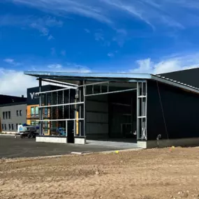 A partially constructed prefabricated steel frame commercial building in Thornton, ID. The structure features exposed steel framing, black metal siding, and wood paneling. The surrounding area includes a parking lot, dirt ground, and a clear blue sky.