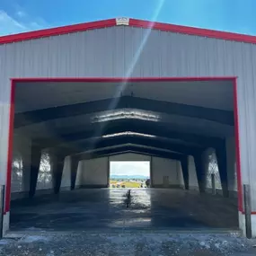 A finished steel structure by Steel Concepts, featuring a gray metal exterior with distinctive red trim. The image showcases the building's large open entrance, revealing a smooth concrete floor and exposed steel beams within. The distant landscape visible through the building highlights the spacious design, suitable for industrial, agricultural, or commercial use. This structure exemplifies Steel Concepts' expertise in delivering high-quality, durable metal buildings.