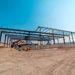 An ongoing steel frame construction project by Steel Concepts, showcasing the initial structural framework. This image captures the early stages of building erection, with visible steel beams, supports, and construction equipment. The concrete foundation and surrounding dirt construction site highlight the progress of this industrial or commercial steel building, demonstrating Steel Concepts' expertise in steel fabrication and construction.