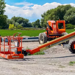 An outdoor image featuring a construction site where Steel Concepts Inc. is operating. In the foreground, a worker wearing a hard hat and camouflage shirt stands near the basket of an orange JLG 660SJ all-terrain boom lift, which is partially visible. Another larger orange piece of heavy equipment is in the background, amidst a gravel surface. Beyond the construction area, there are green fields and trees under a bright blue sky with scattered clouds. The scene conveys a professional constructio