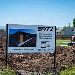 This image features a prominent on-site construction sign for a Steel Concepts project. The sign details the development of a 12,600 square foot Regional Gym in Twin Falls, ID, where Steel Concepts serves as the General Contractor. The sign includes a rendering of the future building, along with Steel Concepts' website and phone number, indicating an active construction site for a retail category facility.