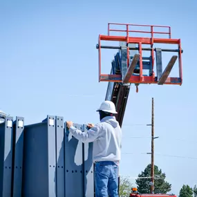 This image, provided by Steel Concepts, illustrates a worker actively engaged in handling substantial steel components on a construction site. The individual, wearing a white hard hat for safety, is shown arranging or securing large, dark gray steel panels. In the background, the lifted forks of a telehandler suggest ongoing material movement and precise placement, highlighting the detailed work involved in steel assembly and construction.