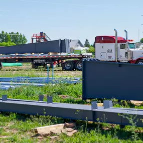 This image, provided by Steel Concepts, illustrates a construction site with significant steel elements. In the foreground, numerous dark gray steel beams are laid out, ready for assembly. A flatbed semi-trailer in the background is loaded with more substantial steel structures, indicative of ongoing material delivery for a large-scale project. This scene highlights the readiness and logistical support for steel-intensive construction.