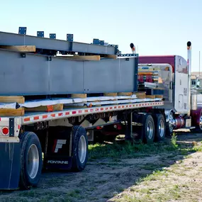 This image, provided by Steel Concepts, captures a crucial moment in construction logistics. A flatbed trailer, bearing substantial dark gray steel beams secured with dunnage, is visible on what appears to be a construction or staging site. A worker, identifiable by their hard hat, is seen gesturing towards the truck, indicating active coordination during the delivery or unloading of these heavy structural components. This scene emphasizes the precision and teamwork involved in handling large-sc